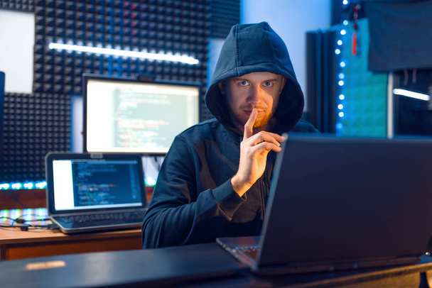 Hacker v kapuci ukazuje palce na svém pracovišti s notebookem a PC, heslem nebo hacknutím účtu. Internetový špion, kriminální životní styl - Fotografie, Obrázek