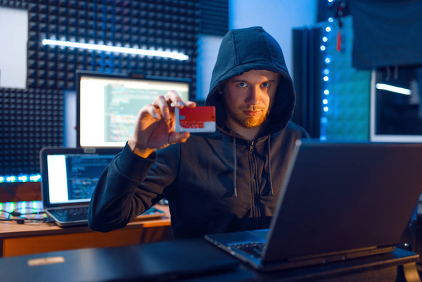 Hacker in hood shows bank credit card at his workplace with laptop and desktop PC, password or finance hacking, darknet user. Internet spy, crime lifestyle, risk job, network criminal - Photo, Image