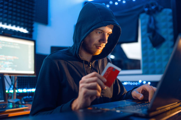 Hacker mit Kapuze zeigt Kreditkarte an seinem Arbeitsplatz mit Laptop und Desktop-PC, Passwort oder Finanz-Hacking, Darknet-Nutzer. Internet-Spion, krimineller Lebensstil, Risiko-Job, Netzwerk-Verbrecher - Foto, Bild