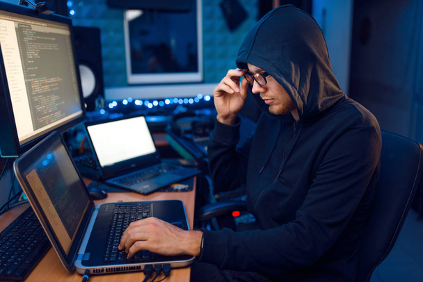 Hacker in hood at his workplace with laptop and desktop PC, website or corporate hacking, darknet user. Internet spy, crime lifestyle, risk job, network criminal - Photo, Image