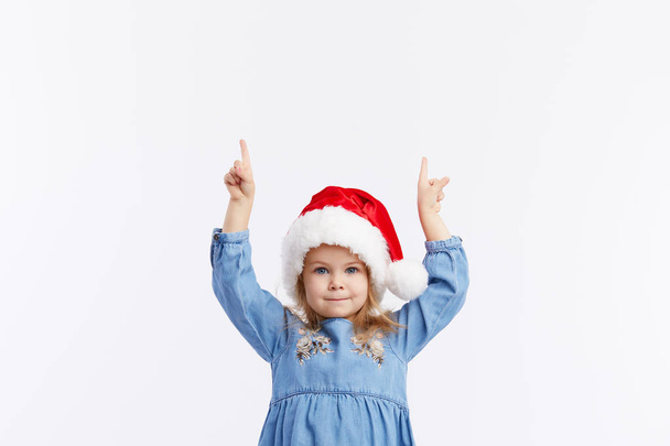 Happy smiling child girl in Santa hat and blue dress having fun on a red isolated background. points with hands up, empty area for text. Merry Christmas. Happy New Year - Φωτογραφία, εικόνα