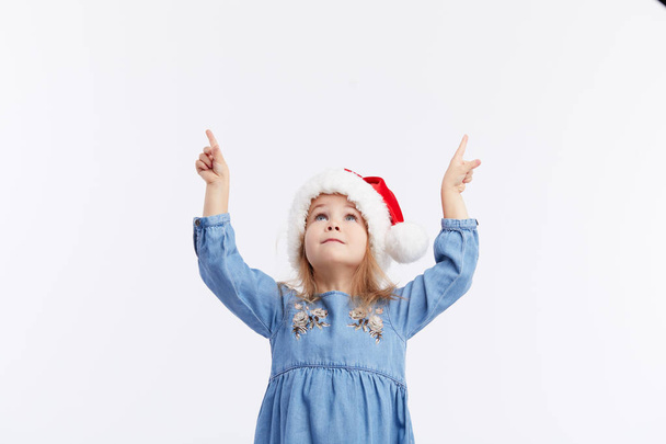 Happy smiling child girl in Santa hat and blue dress having fun on a red isolated background. points with hands up, empty area for text. Merry Christmas. Happy New Year - Photo, Image