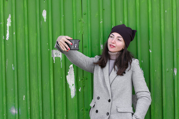 Mädchen macht ein Selfie auf grünem Hintergrund. mit Hut und Mantel. Platz zum Schreiben. gekleidet in einer kalten Pagode. Kleidung und Stil. Natürliche Emotionen - Foto, Bild
