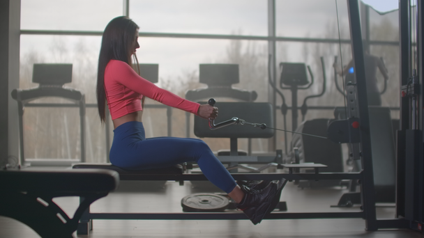 Intensive power training in fitness center. A woman sitting pulls the weight of a simulator against the backdrop of large Windows and treadmills of the gym - Footage, Video