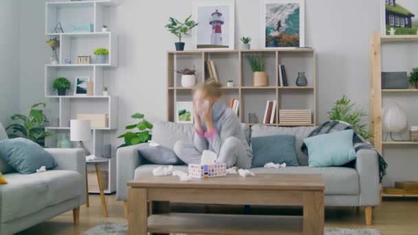 Girl with a cold sneezes while sitting on a couch in a room - Filmmaterial, Video