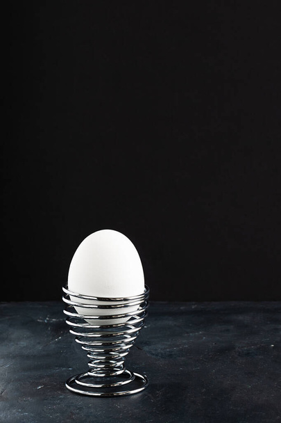 White egg in metal helix egg cup on black chalkboard and empry black background with copy space, Creative concept for breakfast and Easter, Vertical - Fotó, kép