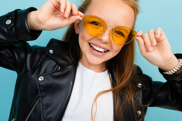 cute little girl in black leather jacket posing with colorful sunglasses against blue background  - Фото, изображение