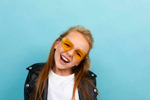 cute little girl in black leather jacket posing with colorful sunglasses against blue background  - Fotó, kép