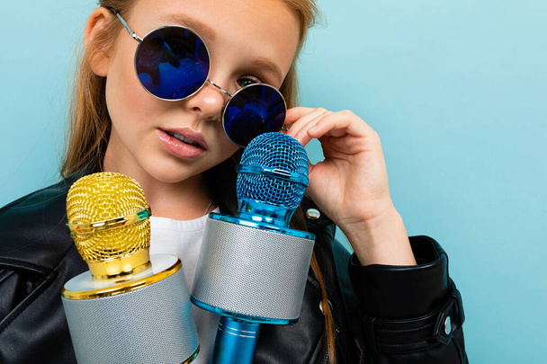 roztomilý holčička v černé kožené bundě pózovat se dvěma mikrofony proti modrému pozadí  - Fotografie, Obrázek
