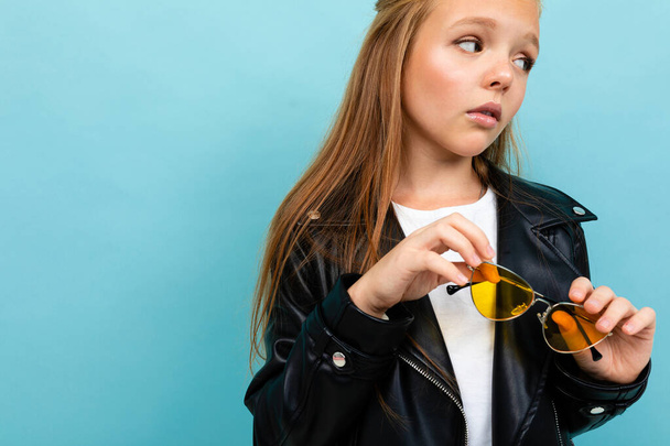 милая маленькая девочка в черной кожаной куртке позирует в красочных солнцезащитных очках на синем фоне
  - Фото, изображение