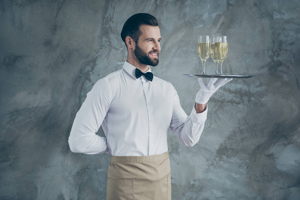 Turned photo of cheerful positive handsome waiter with hand behind his back smiling toothily holding plate with glasses of champagne isolated grey concrete wall color background - Photo, Image