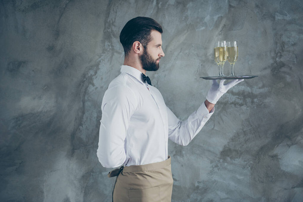 Profile side photo of attractive handsome in apron holding plate with glasses of champagne with bristle isolated grey color concrete wall background - Photo, image
