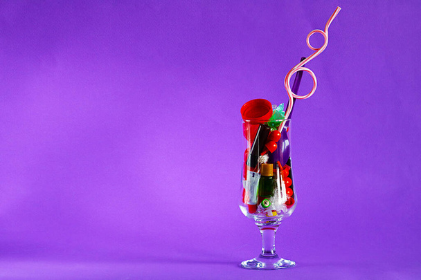 Coctail glass with plastic straw full of plastic and battery waste as imitation of real drink on vertical violet background with copy space. Ecology concept of getting pollution to food, drinks - Foto, immagini