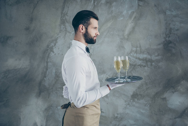 Side profile photo of attractive server holding tray with champagne glasses with bristle isolated grey concrete wall color background - Foto, imagen
