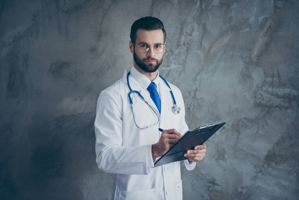 Portrait of concentrated doctor guy hold his clipboard write patients complaints wear white medical coat isolated over grey color background - Фото, изображение