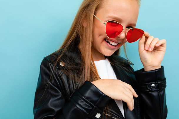 cute little girl in black leather jacket posing with colorful sunglasses against blue background  - Foto, Imagen