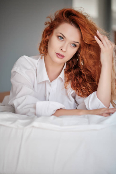 Smiling and looking forward. Hot redhead girl sits on the white  - Фото, зображення