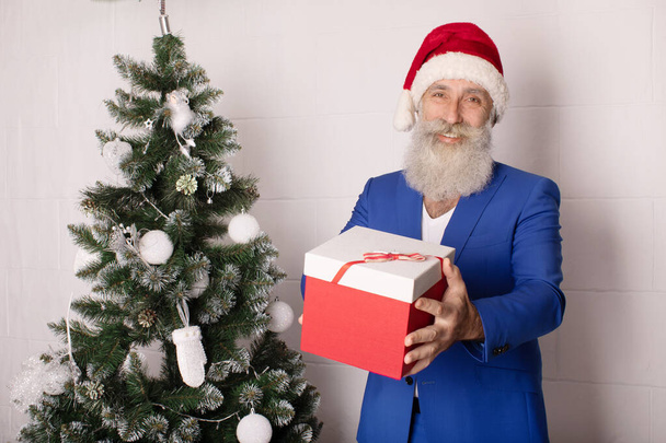 Uśmiechnięty nowoczesny brodaty biznesmen w Santa Hat prezentuje prezent na białym - Zdjęcie, obraz