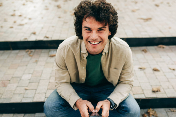 Portrait of smiling young man standing outdoors, typing messages on mobile phone. Young male with curly hair sittiing on the stairs in the city street resting outside, browsing on his smart phone - Fotografie, Obrázek