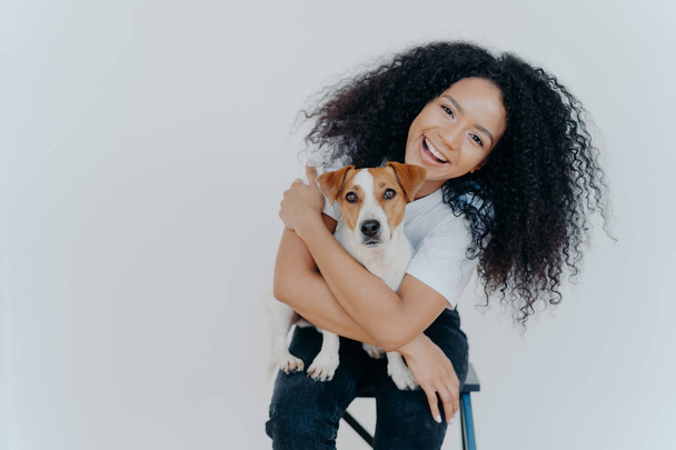 Pleasant looking curly girl tilts head, smiles happily, embraces favourite dog, has good time with pet, wears casual t shirt and jeans, sits on comfortable chair against white background. Relationship - Photo, Image