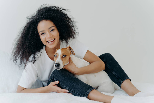Horizontal shot of glad Afro woman rests in bed with dog, have playful mood, pose together in bedroom against white background. Girl relaxes at home with jack russell terrier. Sweet funny moment - Foto, Imagem