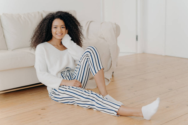Ethnic girl feels relaxed and satisfied, sits on floor near comfortable sofa in empty room, wears white sweater, striped pants and socks, enjoys domestic atmosphere, enjoys coziness and comfort - Photo, Image