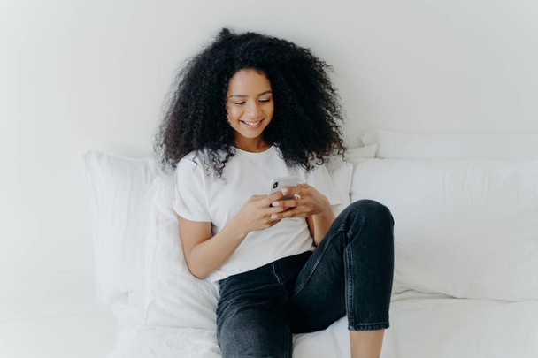Relajada hermosa mujer Afro con expresión alegre utiliza el teléfono móvil moderno, navega por Internet, posa en una cama cómoda blanca, conectado a wifi, navega por la página web, disfruta de la atmósfera doméstica
 - Foto, Imagen