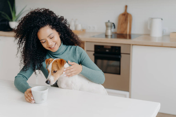 Happy Afro African woman with curly hairstyle treats dog in kitchen, pose at white table with mug of drink, enjoy domestic atmosphere, have breakfast together. People, animals, home concept. - Photo, Image