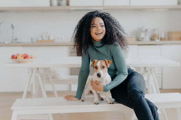 Joyful Afro woman sits at white bench together with dog against kitchen interior, table with plate full of red apples, get pleasure while playing at home. Animal owner feels care and responsibility - Foto, immagini