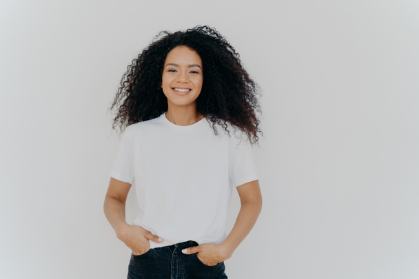 Isolated shot of young African American woman wears white t shirt, expresses good emotions, stands alone indoor, poses for photo, has casual talk with friend, enjoys free time. People and happiness - 写真・画像