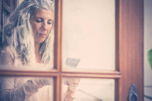 Beautiful vintage old style portrait of alternative senior woman typing and reading a mobile phone device at home viewed from the window  - Photo, Image