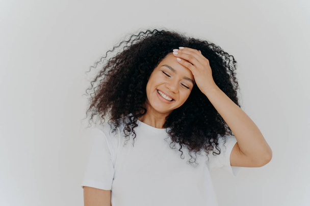 Photo of happy lady keeps hand on forehead, tilts head, smiles joyfully, keeps eyes closed, has curly Afro hair, wears casual white t shirt, enjoys pleasant moment in life, feels little bit shy - Photo, Image