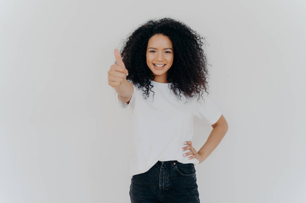 Photo of happy curly woman with crisp hair raises thumb up, gives approval, says sounds good, makes supportive gesture, keeps other hand on waist, dressed casually, isolated over white background - 写真・画像