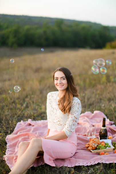 Jeune fille caucsienne assise sur plaid avec bouteille de vin et de fruits, bulles de savon
. - Photo, image