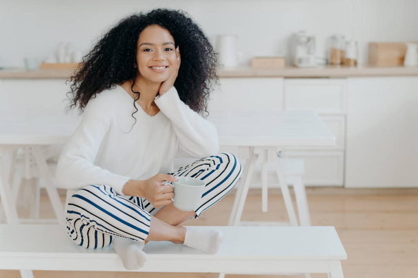 Indoor shot of pretty African American woman wears white jumper, striped pants, socks, poses on bench with cup of tea spend leisure at home in cozy kitchen feel relaxed. Mujer disfruta del café de la mañana
 - Foto, imagen