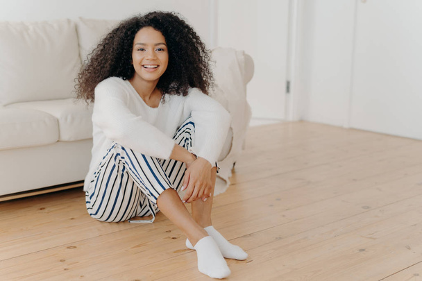 Indoor shot of restful happy African American woman sits on floor in living room, white sofa in background, wears fashionable clothes and socks, expresses positive emorions, rests at home alone - Фото, зображення