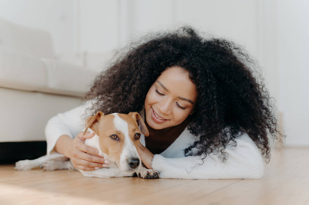 Relaxed Afro woman with crisp dark hair lies on floor, plays with cute puppy, has fun with jack russell terrier dog wears white sweater being in living room. Happy owner petting lovely domestic animal - Photo, Image