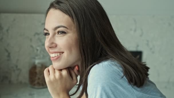 Side view of pretty happy young brunette woman smiling and looking at the window while sitting at the kitchen - Video, Çekim