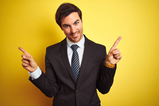 Giovane uomo d'affari bello indossa vestito e cravatta in piedi su sfondo giallo isolato sorridente fiducioso punta con le dita in direzioni diverse. Copia spazio per pubblicità
 - Foto, immagini