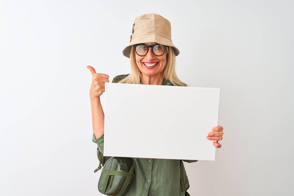 Mujer senderista mayor con sombrero de cantimplora sosteniendo pancarta sobre fondo blanco aislado feliz con una gran sonrisa haciendo signo de ok, pulgar hacia arriba con los dedos, señal excelente
 - Foto, imagen