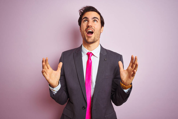 Young handsome businessman wearing suit and tie standing over isolated pink background crazy and mad shouting and yelling with aggressive expression and arms raised. Frustration concept. - Fotó, kép