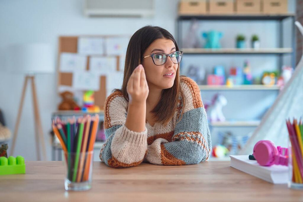 Jeune belle prof femme portant pull et lunettes assis sur le bureau à la maternelle Faire geste italien avec la main et les doigts expression confiante
 - Photo, image