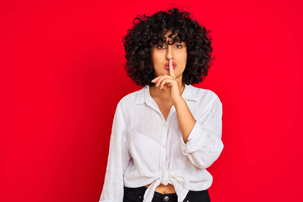 白いカジュアルなシャツを着た巻き毛の若いアラブ人女性は、唇に指で静かにするように求めて孤立した赤い背景の上に。沈黙と秘密の概念. - 写真・画像