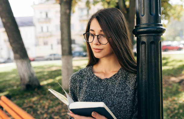 Mulher muito jovem vestindo suéter e óculos transparentes, de pé ao ar livre na rua da cidade e lendo o livro enquanto espera seus amigos. Jovem estudante aprendendo na rua da cidade
.  - Foto, Imagem