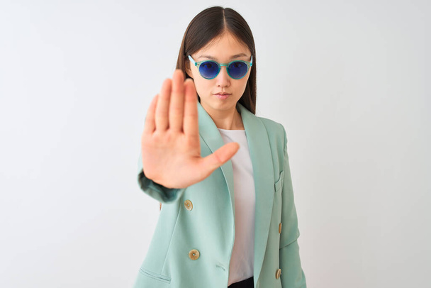 Empresaria china con chaqueta y gafas de sol sobre fondo blanco aislado con la mano abierta haciendo stop sign con expresión seria y segura, gesto de defensa
 - Foto, imagen