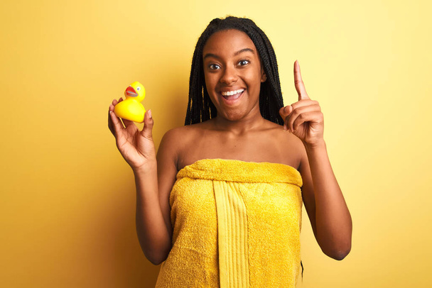 Afrikai amerikai nő visel zuhany törölköző tartó játék kacsa felett elszigetelt sárga háttér meglepett egy ötlet vagy kérdés ujjal mutogató boldog arc, első számú - Fotó, kép