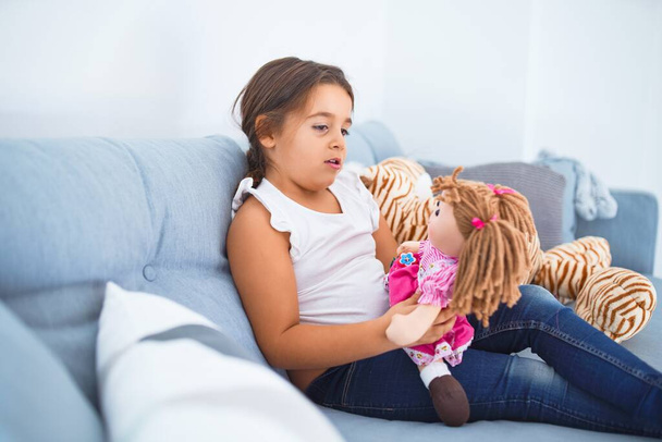Criança adorável sorrindo feliz. Sentado no sofá abraçando boneca em casa
 - Foto, Imagem