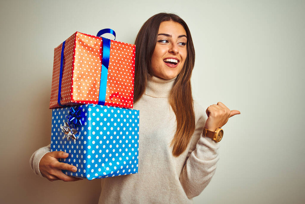 Young beautiful woman holding birthday gifts standing over isolated white background pointing and showing with thumb up to the side with happy face smiling - 写真・画像