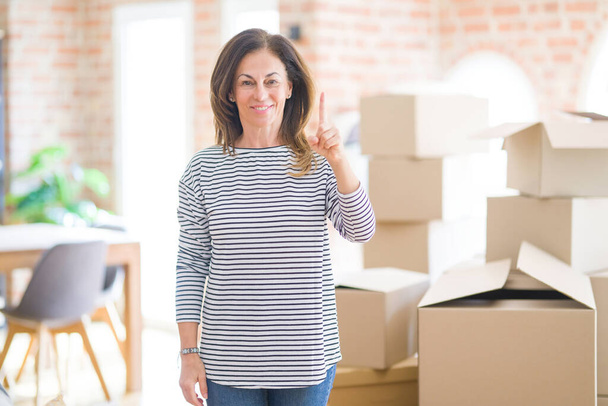 Женщина средних лет переезжает в новый дом вокруг картонных коробок показывая пальцем вверх с успешной идеей. Взволнован и счастлив. Номер один
. - Фото, изображение