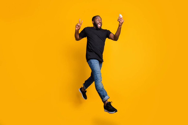 Full size photo of funny crazy afro american guy have journey make v-sign selfie go walk record video smartphone jump wear t-shirt denim jeans isolated yellow color background - Foto, Bild